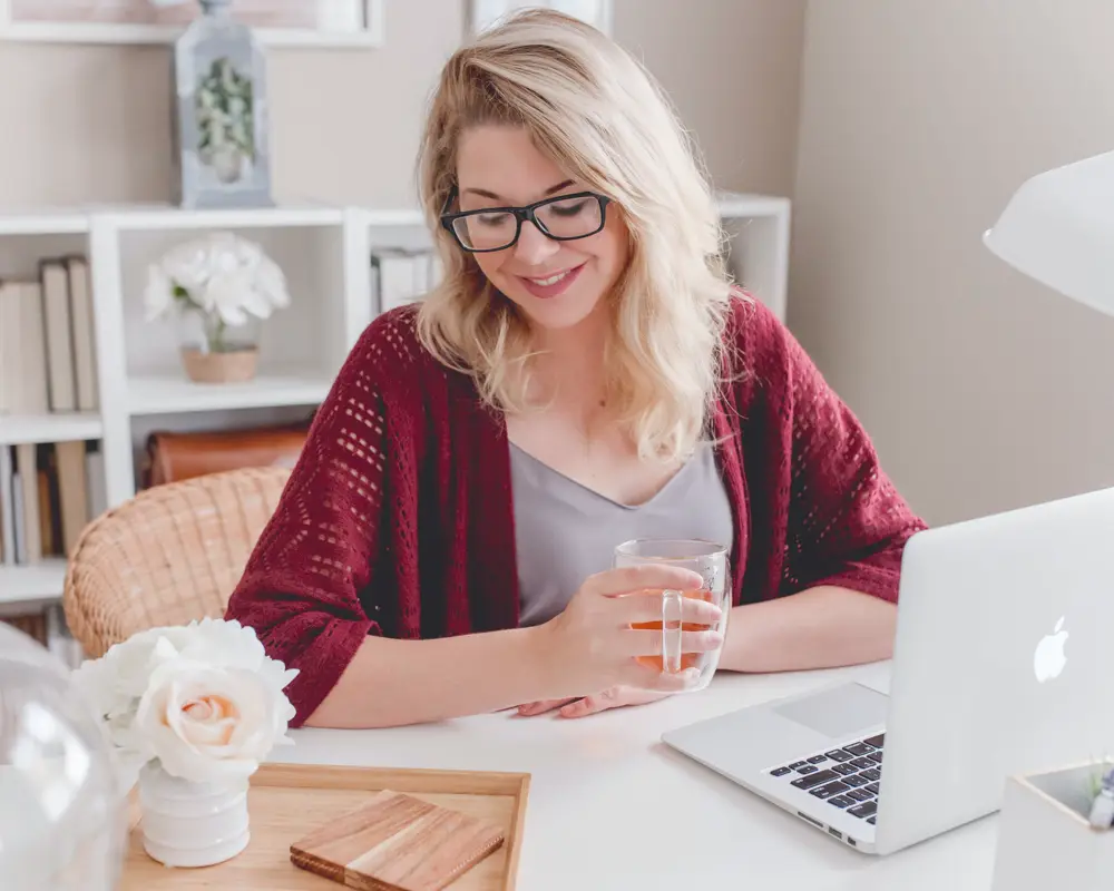 A smiling woman at her desk, mug in hand, in front of her laptop